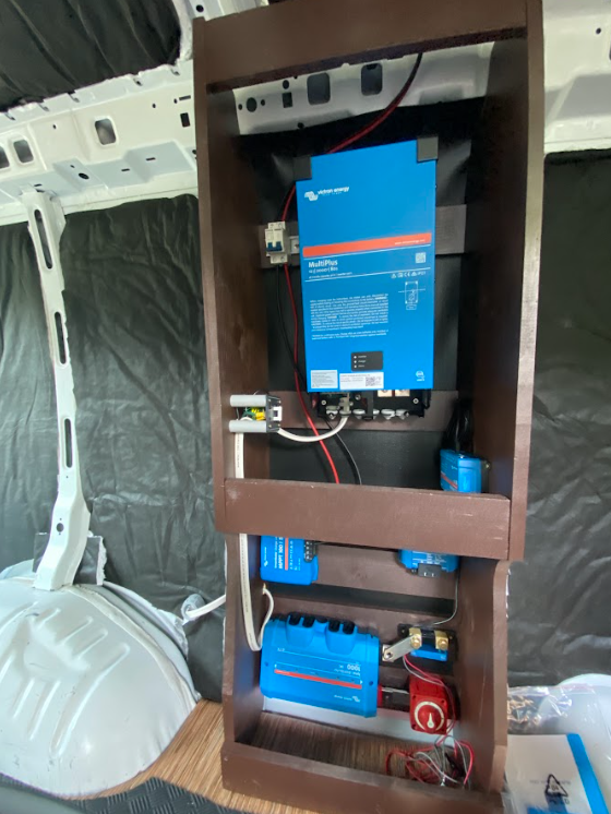 The wooden cabinet frame, bolted to the inside of the cargo van and holding some blue electrical devices which have been mounted to the inside.