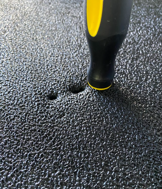 A black mat with three holes in it; one of the holes also has a screwdriver inserted to the hilt.