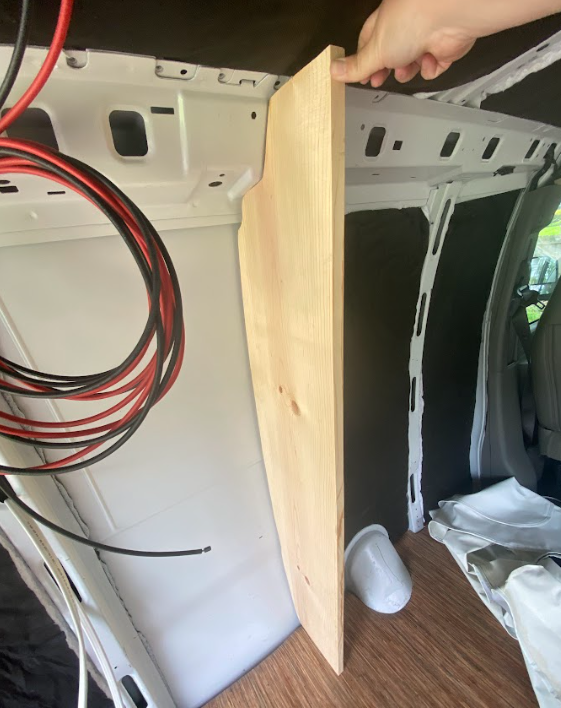 A board being held perpendicular and vertical against the inside of a cargo van. The board has been cut to follow the contours of the van wall.